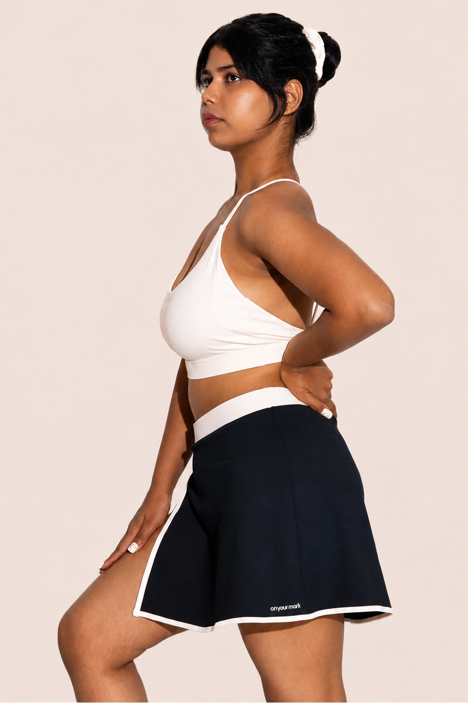 A woman posing in a side profile wearing a white sports bra and a navy blue athletic skirt with white trim, set against a solid light beige background.